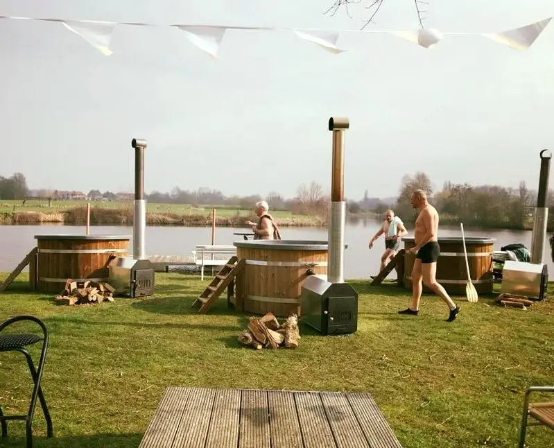 Mannen lopen langs een houten buitenbarbecueplek aan een meer, met houtblokken en gasbarbecues op gras.